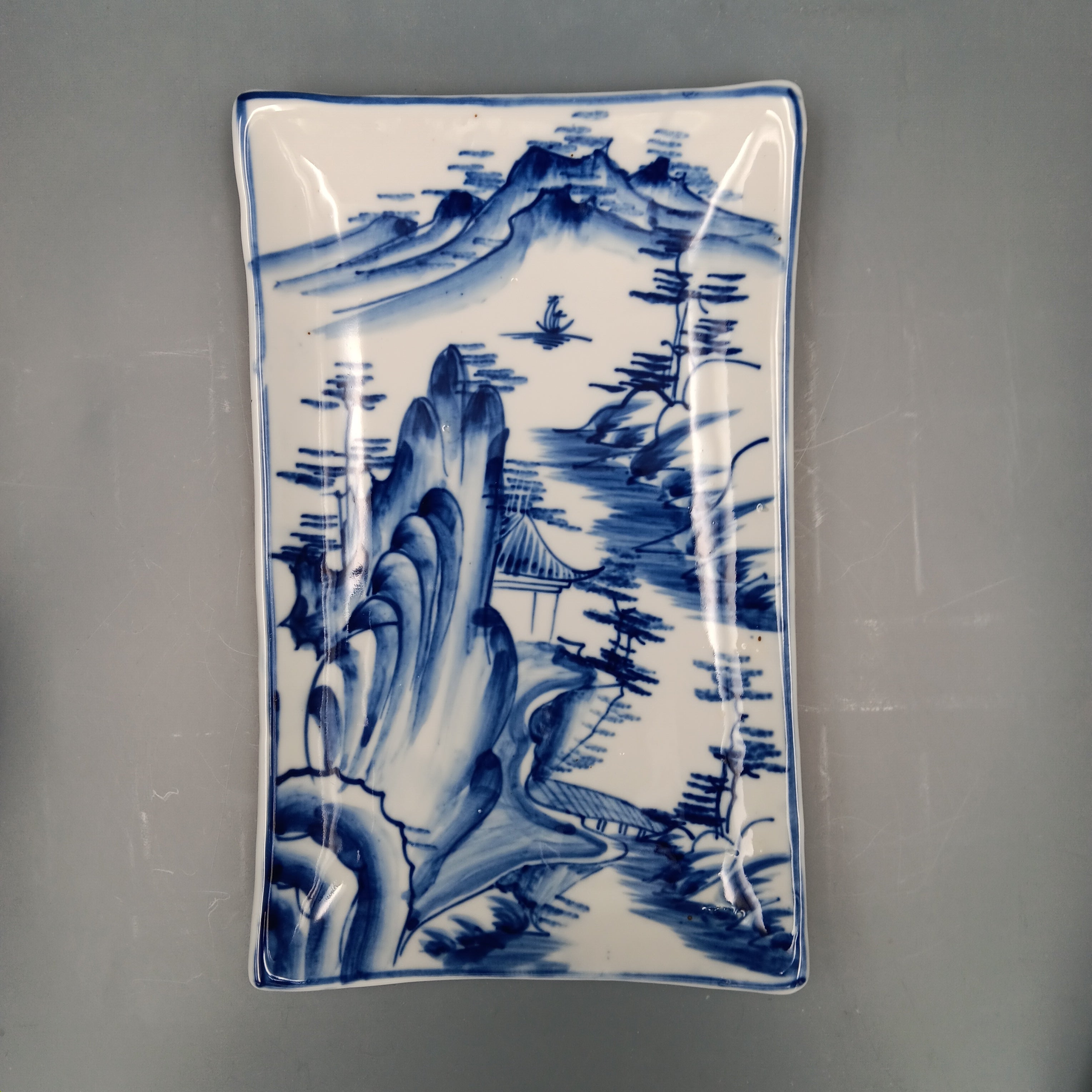 Antique Chinese blue and white porcelain plate with hand-painted landscape scene featuring mountains, trees, and a pavilion in cobalt blue on a rectangular ceramic dish.