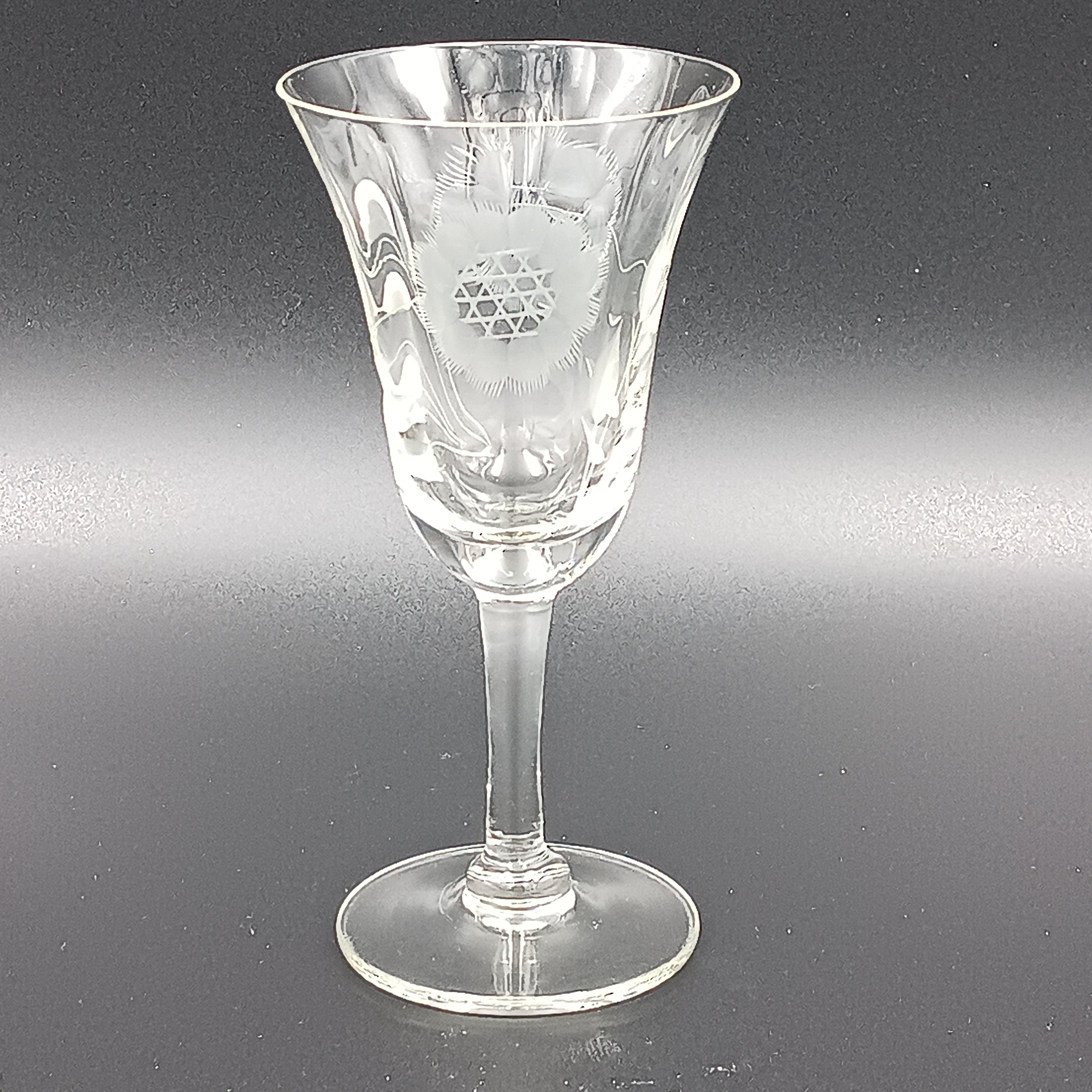 Vintage John Hughes cut crystal wine glass, featuring the iconic Corn Flower pattern, shown lying on its side against a dark reflective surface. The flared bowl displays intricate floral etching, with a slender stem and flat circular base