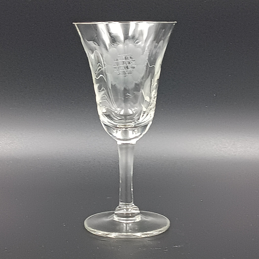 Vintage John Hughes cut crystal wine glass, featuring the iconic Corn Flower pattern, shown lying on its side against a dark reflective surface. The flared bowl displays intricate floral etching, with a slender stem and flat circular base