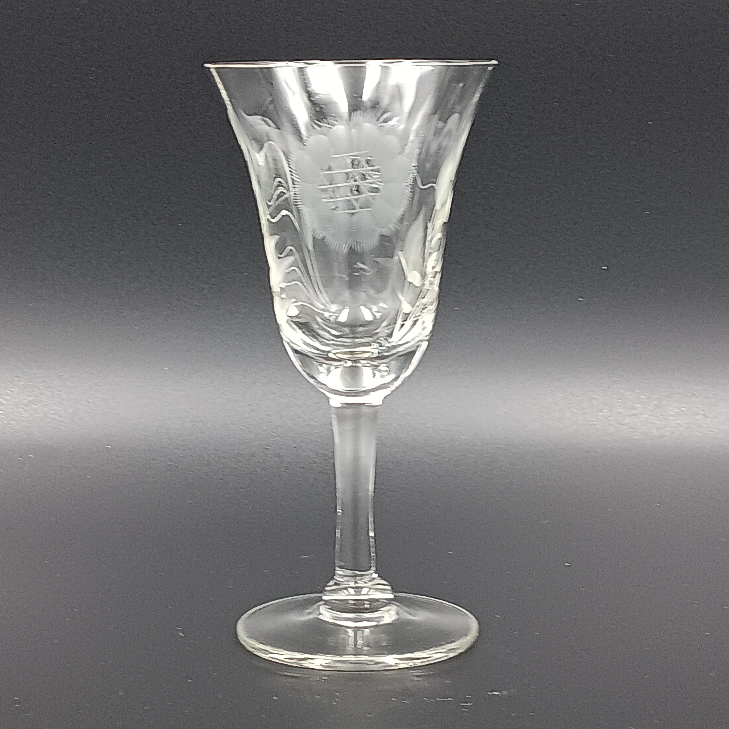 Vintage John Hughes cut crystal wine glass, featuring the iconic Corn Flower pattern, shown lying on its side against a dark reflective surface. The flared bowl displays intricate floral etching, with a slender stem and flat circular base