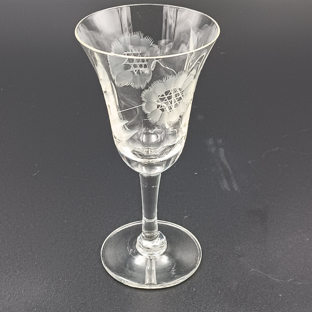 Vintage John Hughes cut crystal wine glass, featuring the iconic Corn Flower pattern, shown lying on its side against a dark reflective surface. The flared bowl displays intricate floral etching, with a slender stem and flat circular base