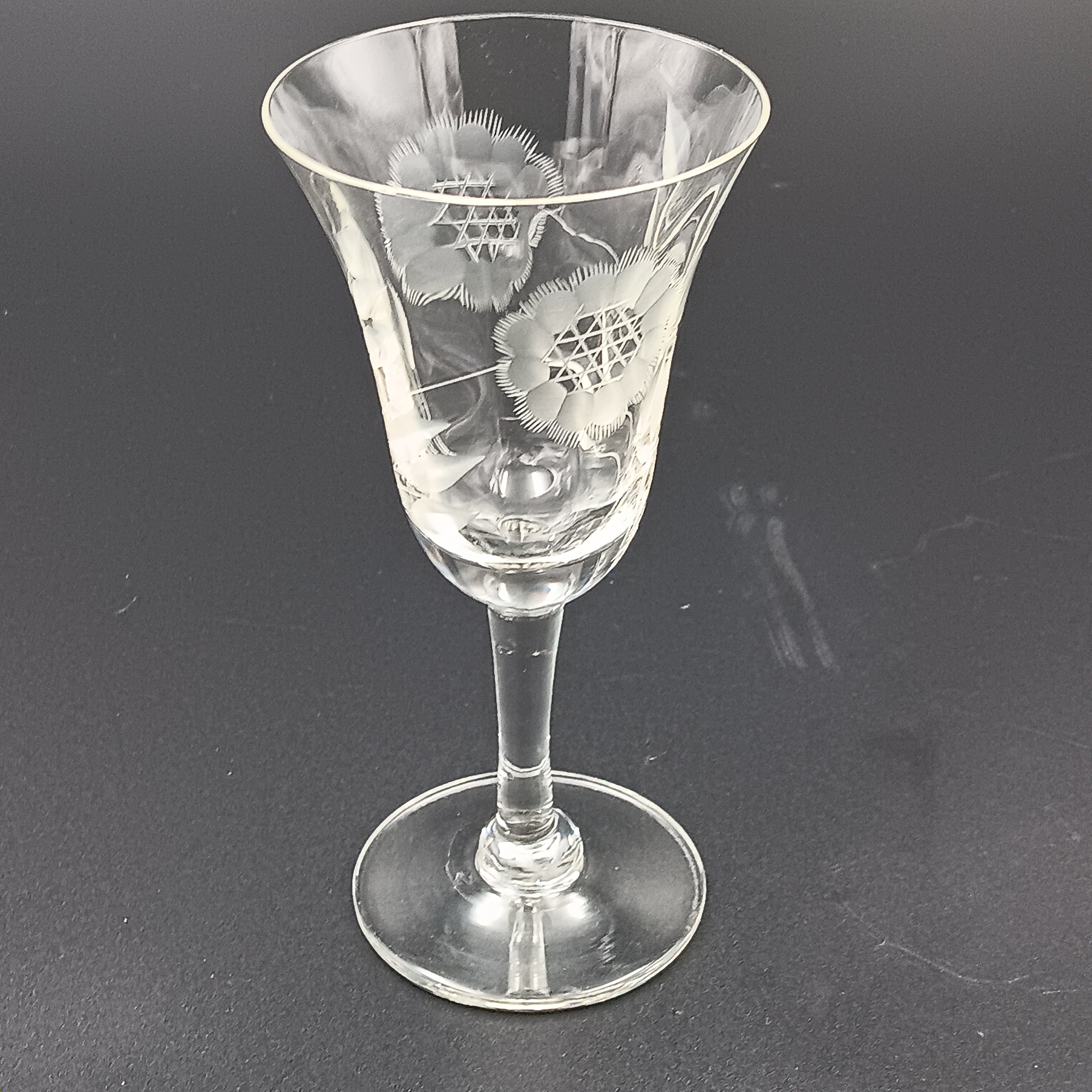 Vintage John Hughes cut crystal wine glass, featuring the iconic Corn Flower pattern, shown lying on its side against a dark reflective surface. The flared bowl displays intricate floral etching, with a slender stem and flat circular base