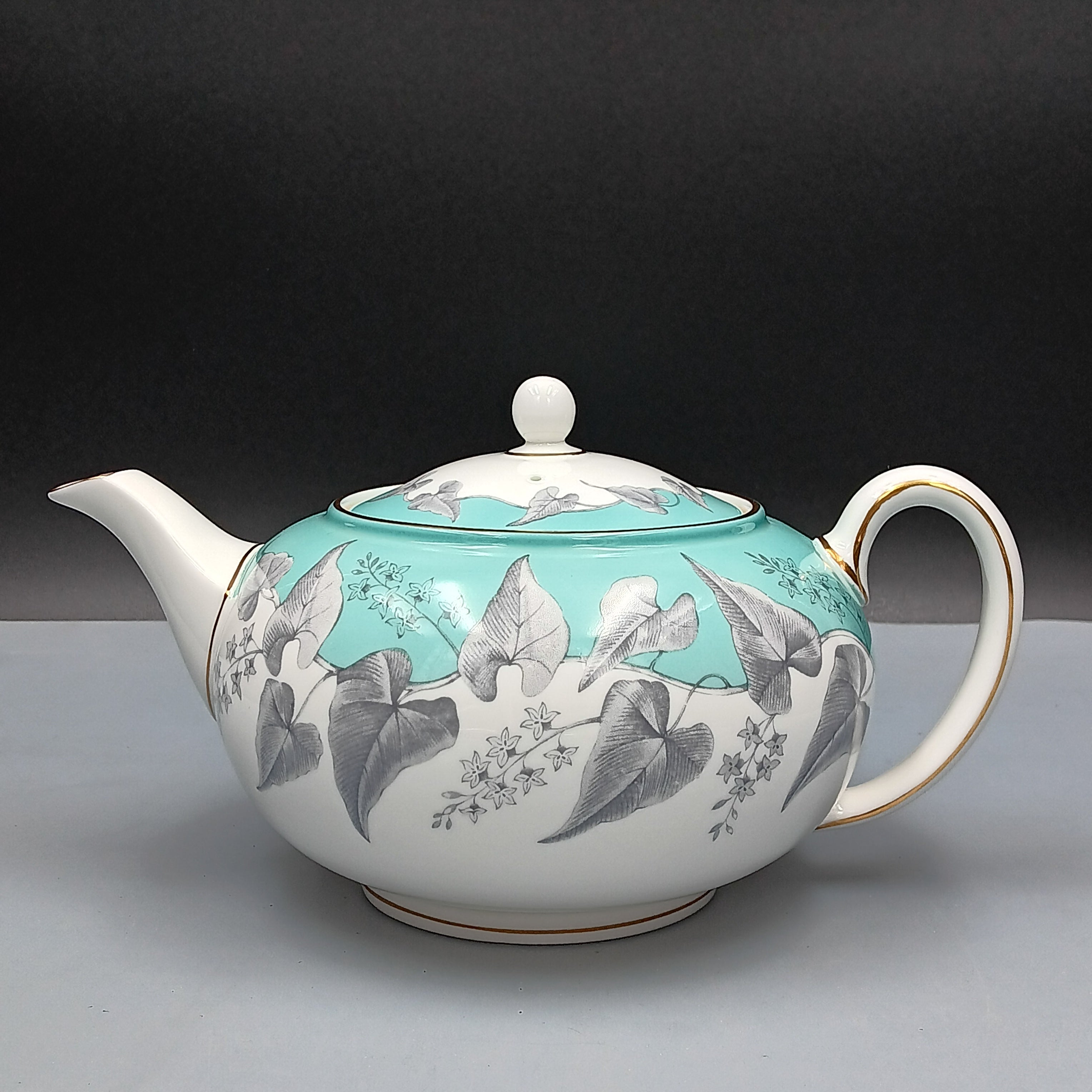 Wedgwood, Buxton, white porcelain teapot with a turquoise floral band, gold-accented handle and spout, and matching lid, displayed against a two-tone background.