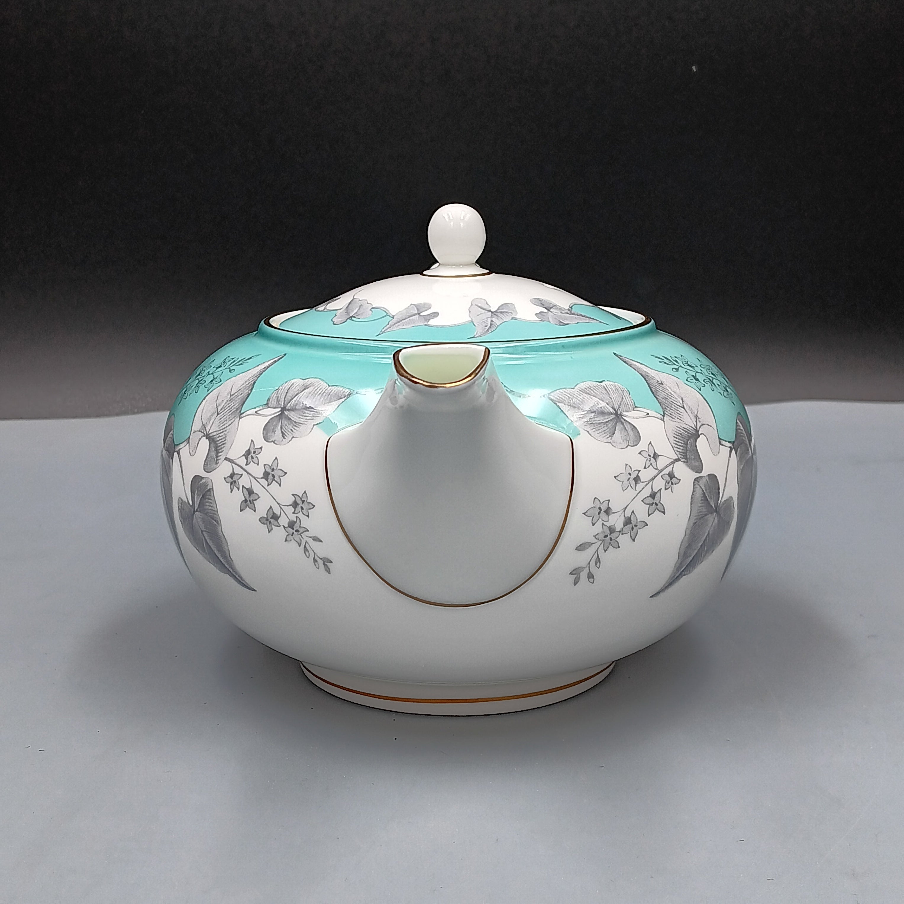 Wedgwood, Buxton, white porcelain teapot with a turquoise floral band, gold-accented handle and spout, and matching lid, displayed against a two-tone background.