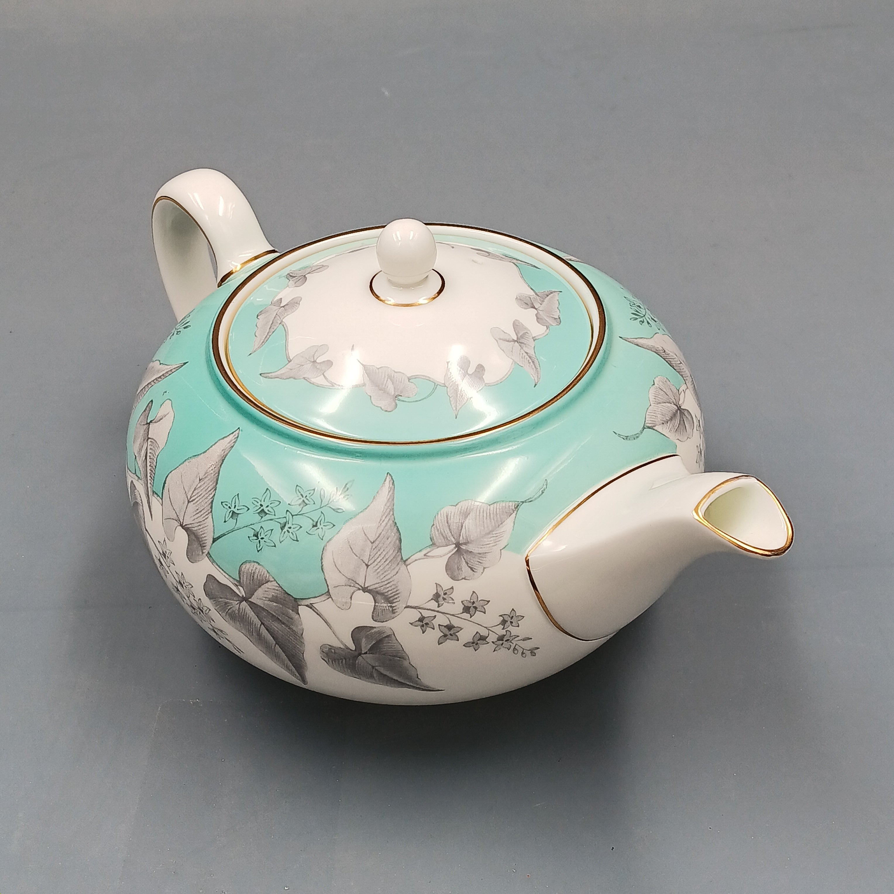 Wedgwood, Buxton, white porcelain teapot with a turquoise floral band, gold-accented handle and spout, and matching lid, displayed against a two-tone background.