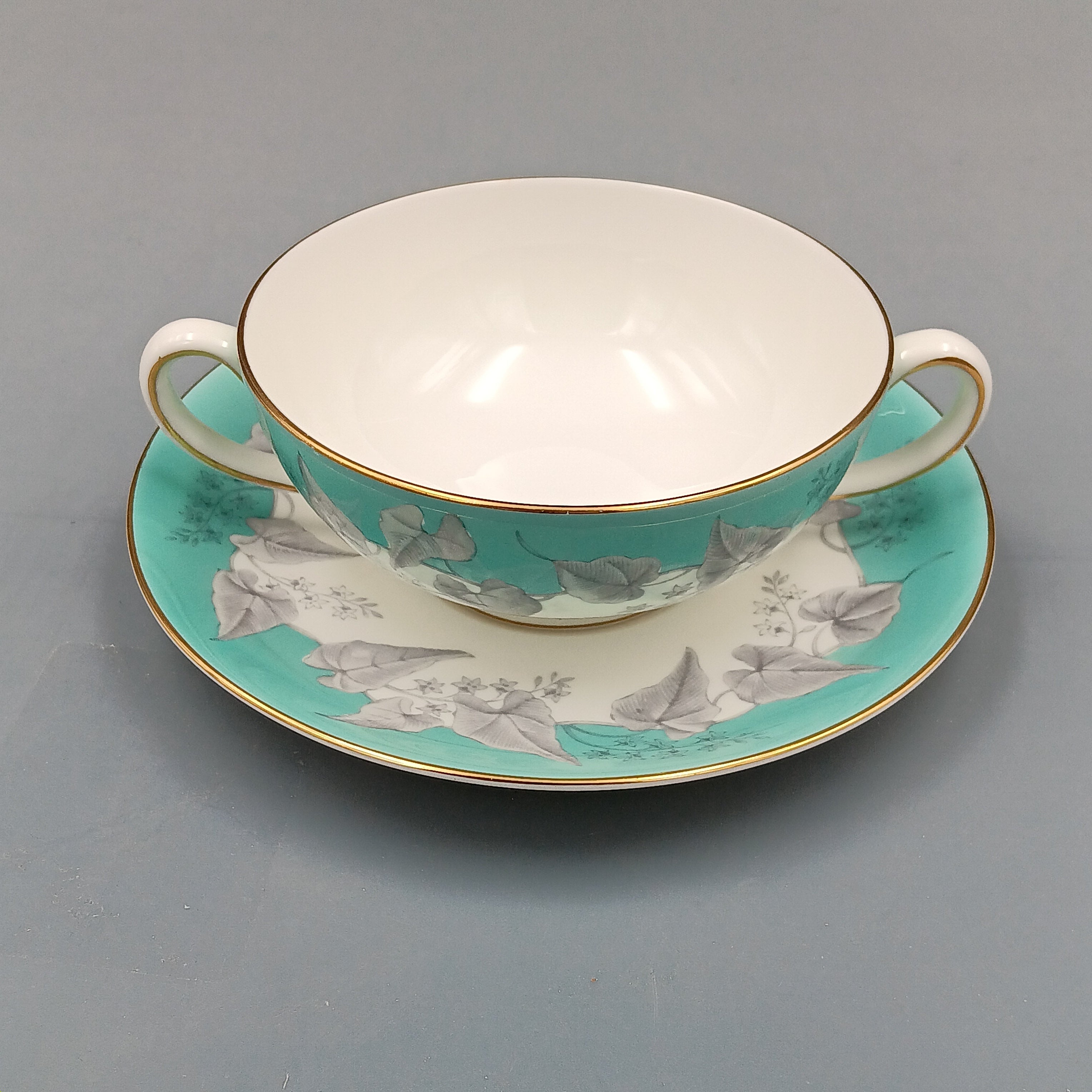 Wedgwood 'Buxton' double-handled soup bowl and saucer set featuring a turquoise background with gray and white floral design, accented by gold trim on edges; elegant English porcelain tableware ideal for collectors.
