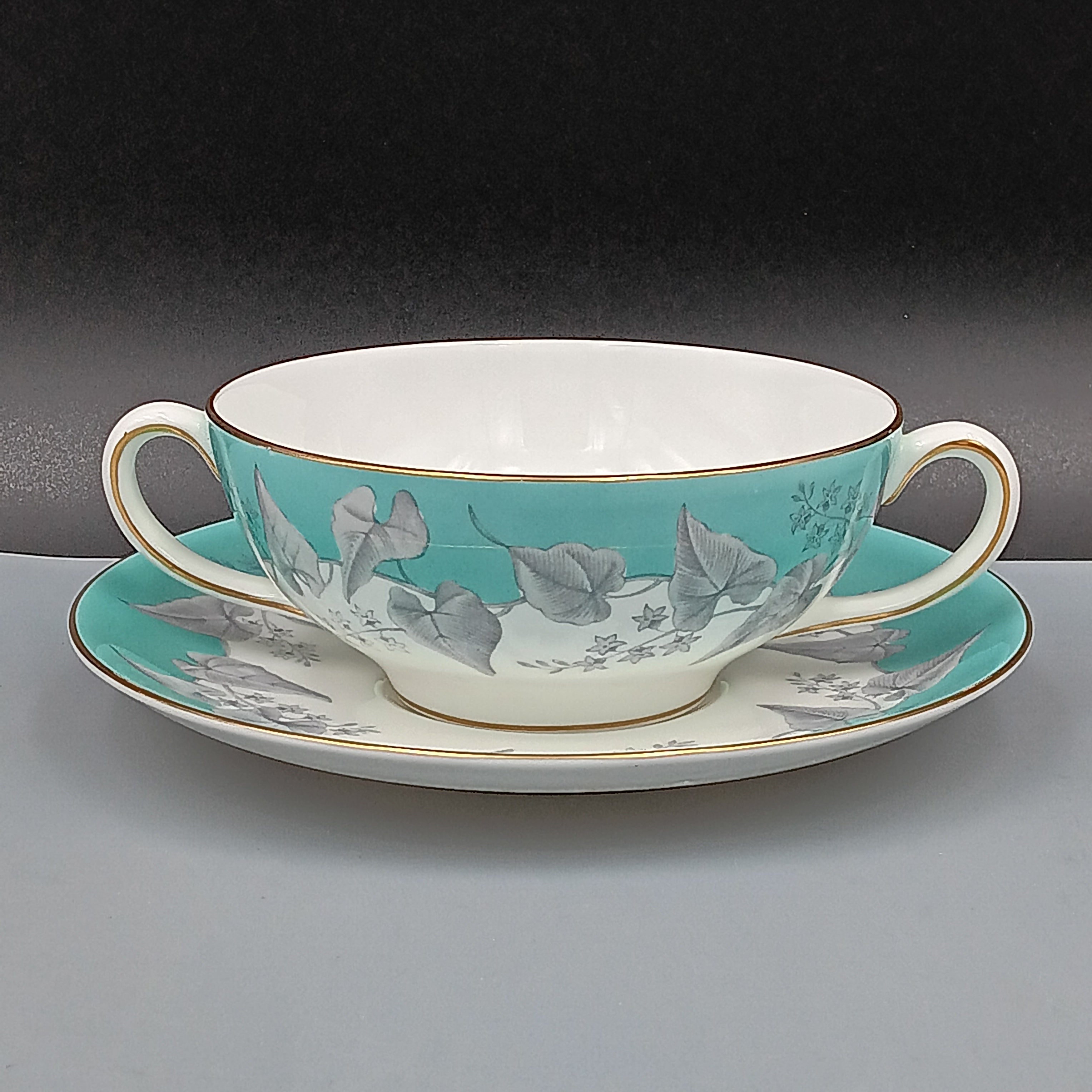 Wedgwood 'Buxton' double-handled soup bowl and saucer set featuring a turquoise background with gray and white floral design, accented by gold trim on edges; elegant English porcelain tableware ideal for collectors.