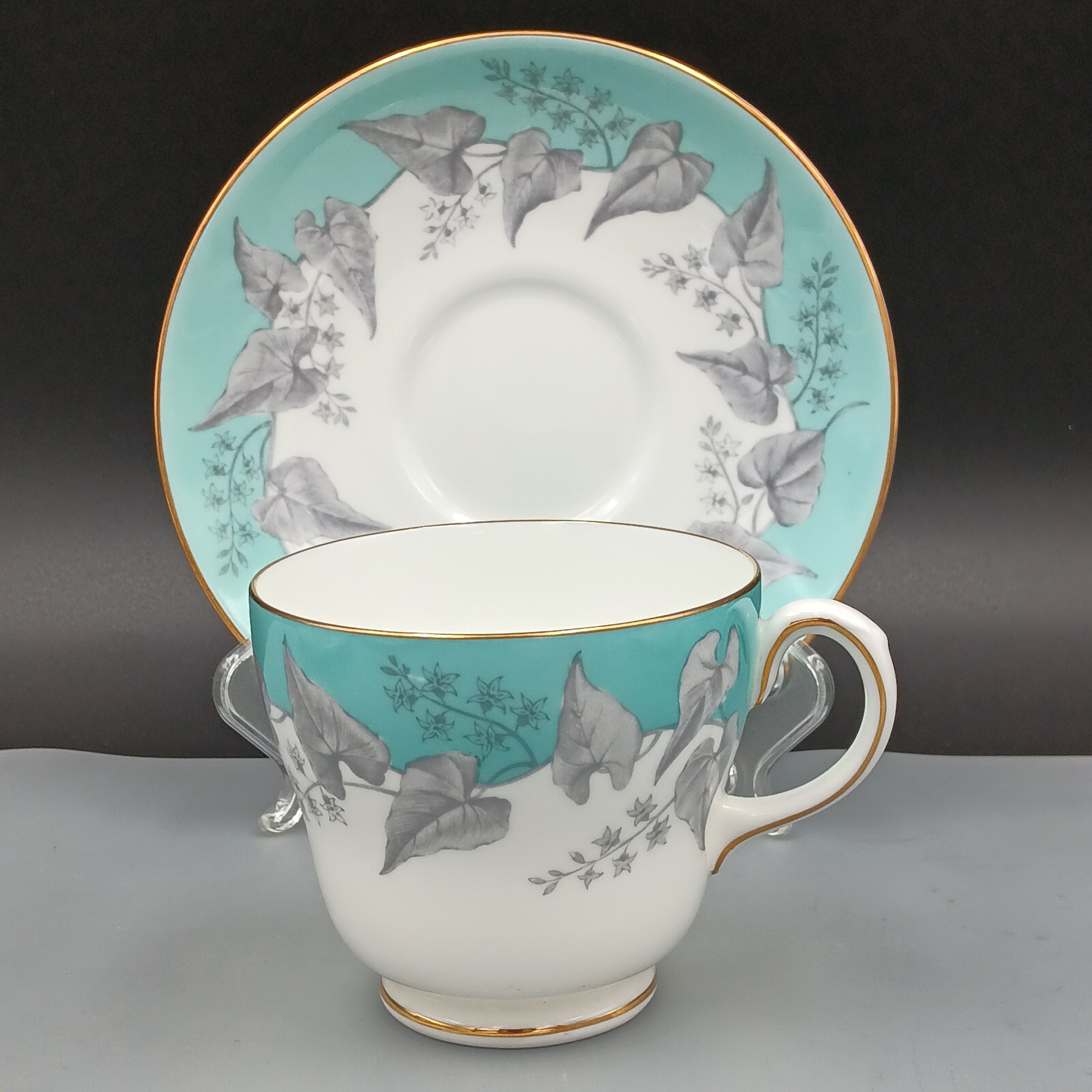 Vintage Wedgwood Buxton Turquoise bone china teacup and saucer set with floral gray leaf pattern, turquoise rim band, and gold trim, displayed upright and flat against a gradient background.